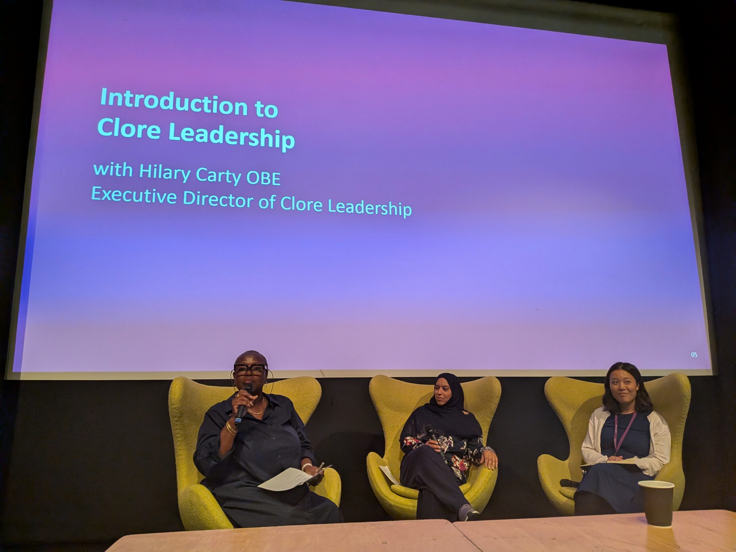 Speakers seated on stage during a discussion about leadership in the cultural sector, with a Clore Leadership presentation projected behind them.